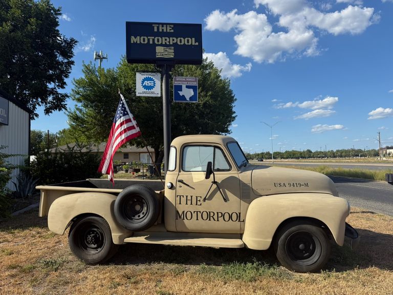 The Motorpool’s vintage tan pickup truck, lettered “The Motorpool,” parked on the grass under The Motorpool sign with an American flag, ASE Certified and Texas vehicle inspection station signs at the Full Service shop in San Antonio